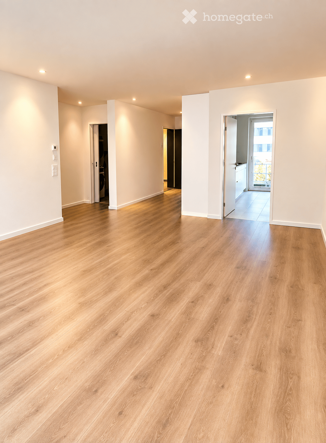 Empty room with light wooden flooring and white walls, featuring recessed lighting and open doorways leading to other rooms.