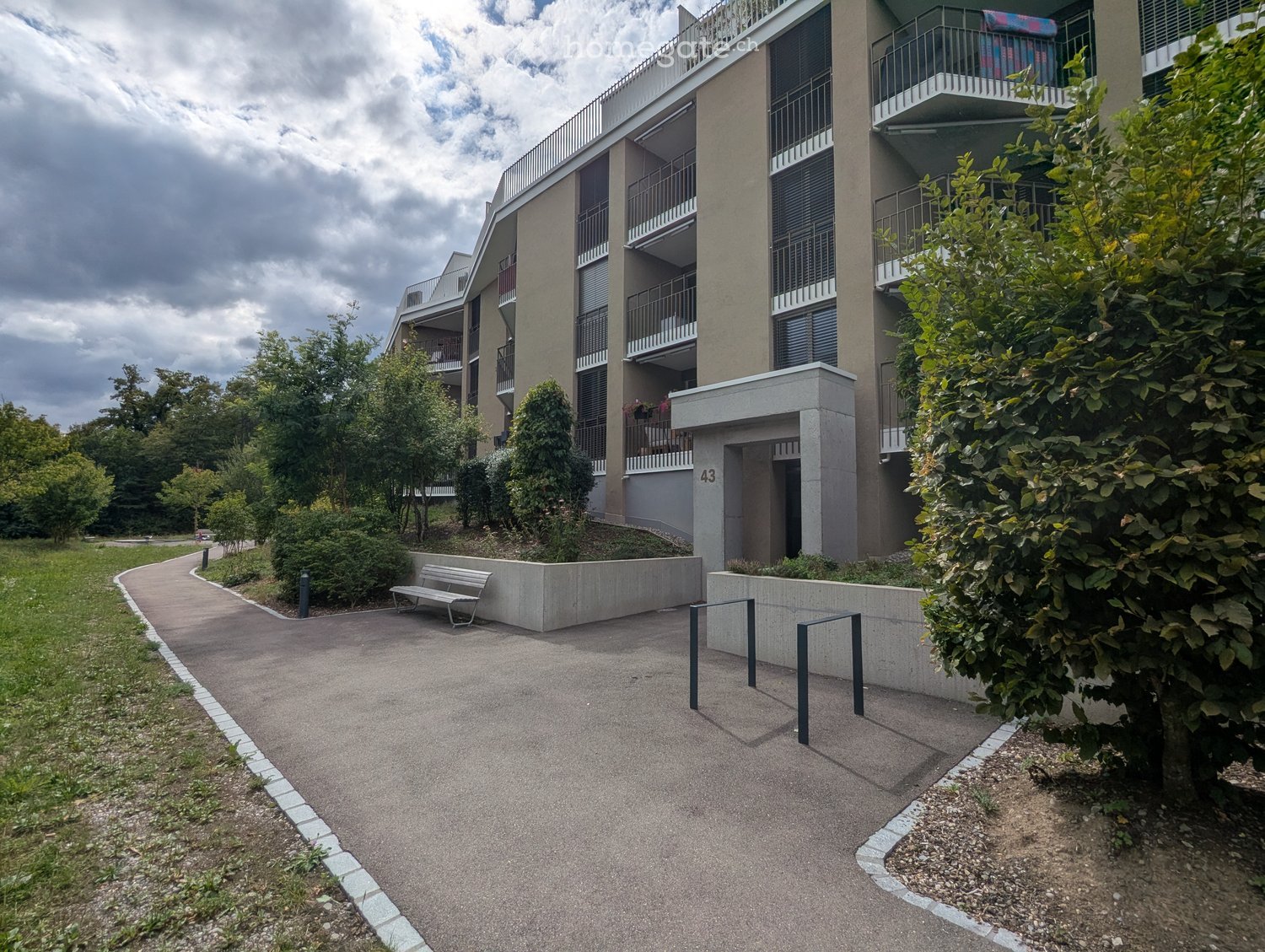 Modern multi-story residential building with balconies, surrounded by greenery and a paved walkway.