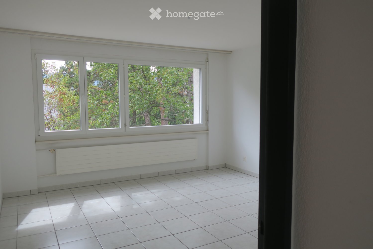 Empty room with white tiled flooring, white walls, and large windows with white frames. A white radiator is mounted below the windows.