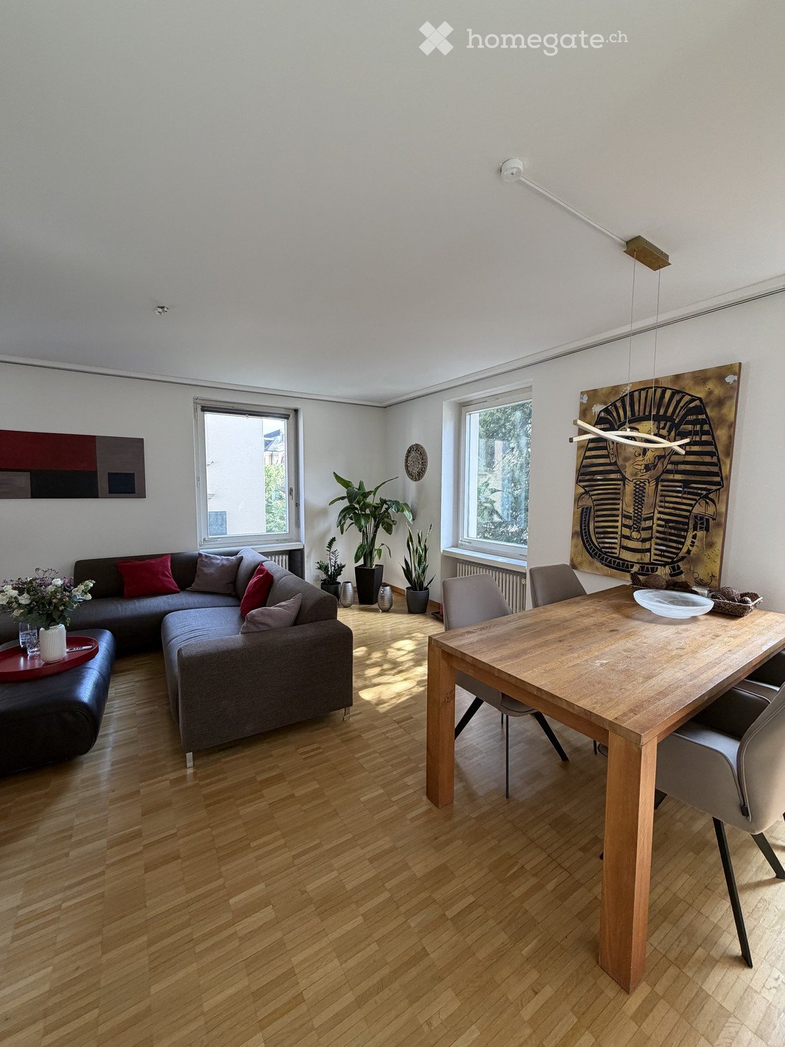 Modern living and dining area with wooden flooring, large windows, grey sectional sofa, wooden dining table, and decorative artwork.