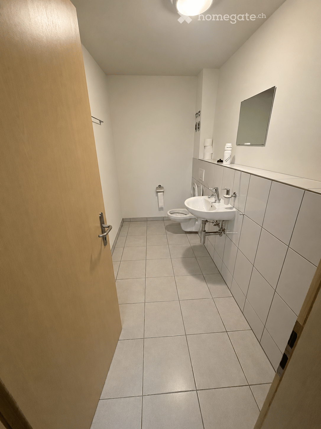 Modern bathroom with white tiled walls and floor, a toilet, a sink with a faucet, a mirror, and a bottle on the counter.