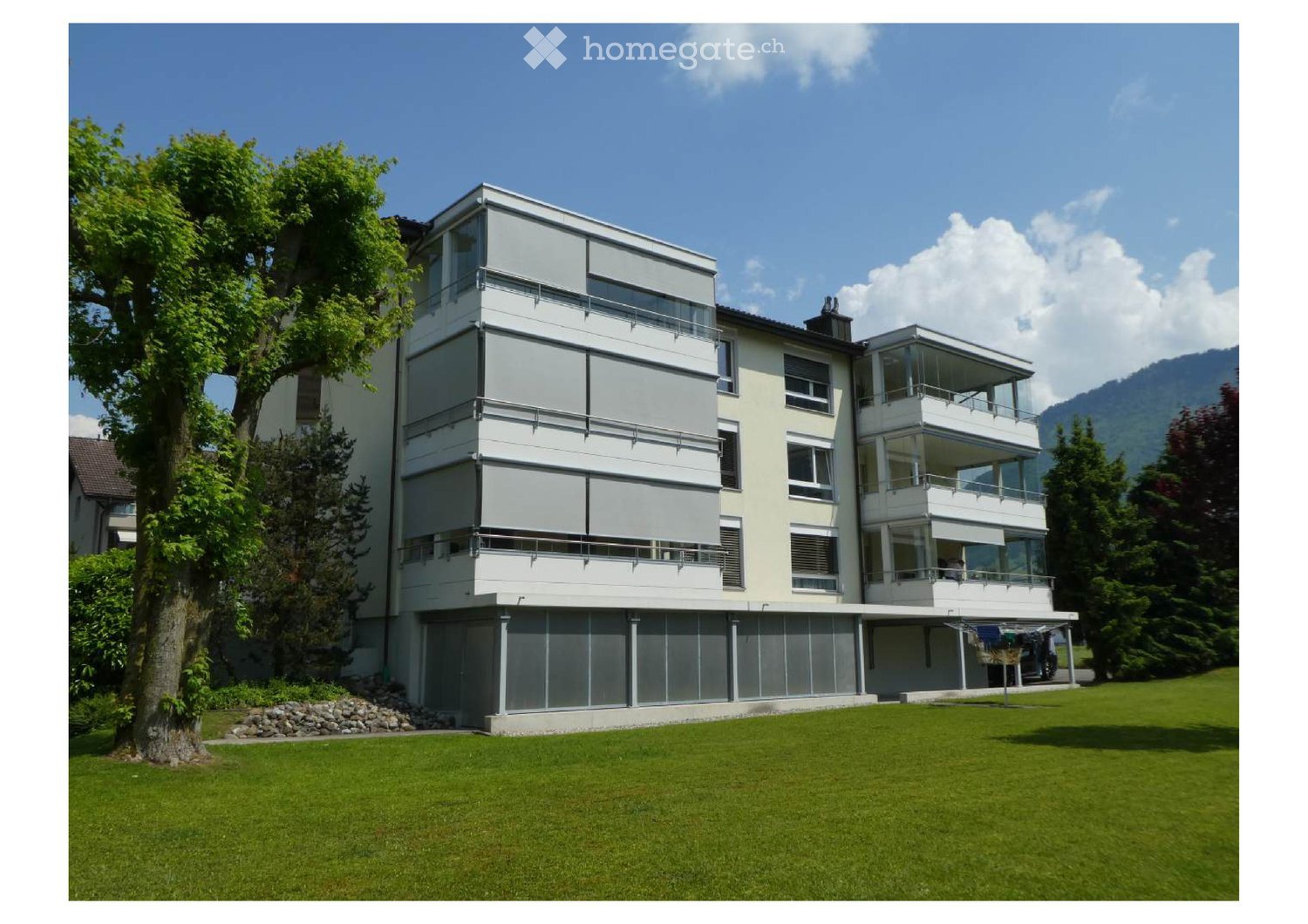 Modern multi-story house with large windows, balconies, and a raised ground floor, surrounded by a well-maintained lawn and trees.