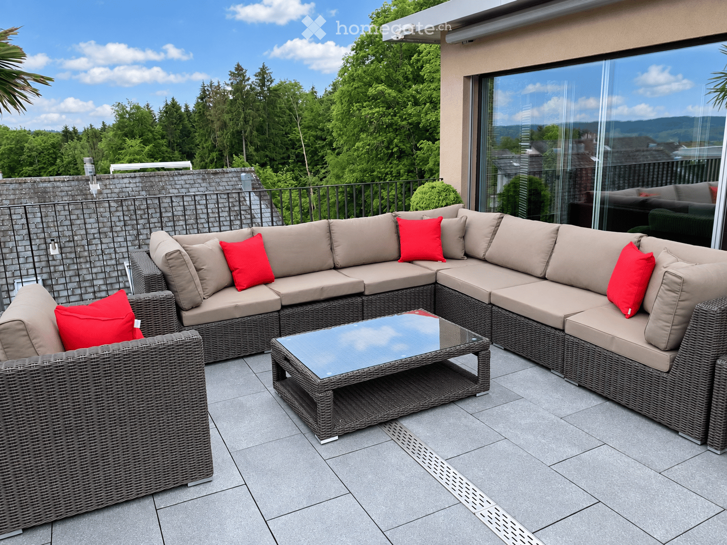 Outdoor patio with a modern wicker sectional sofa, red throw pillows, and a glass coffee table, overlooking a lush green landscape.