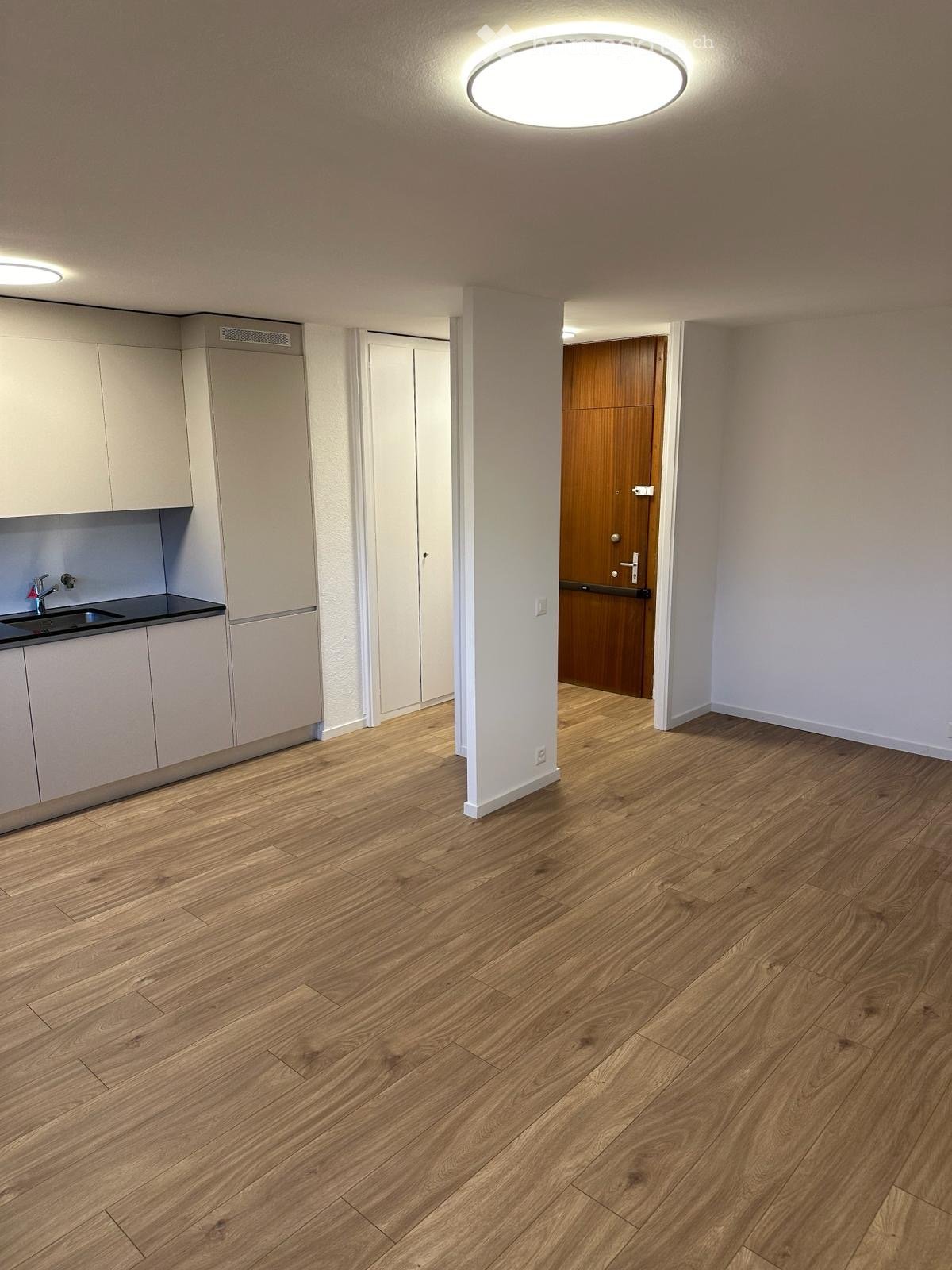 Empty room with wooden flooring, white walls, and a small kitchen area with cabinets and a sink.