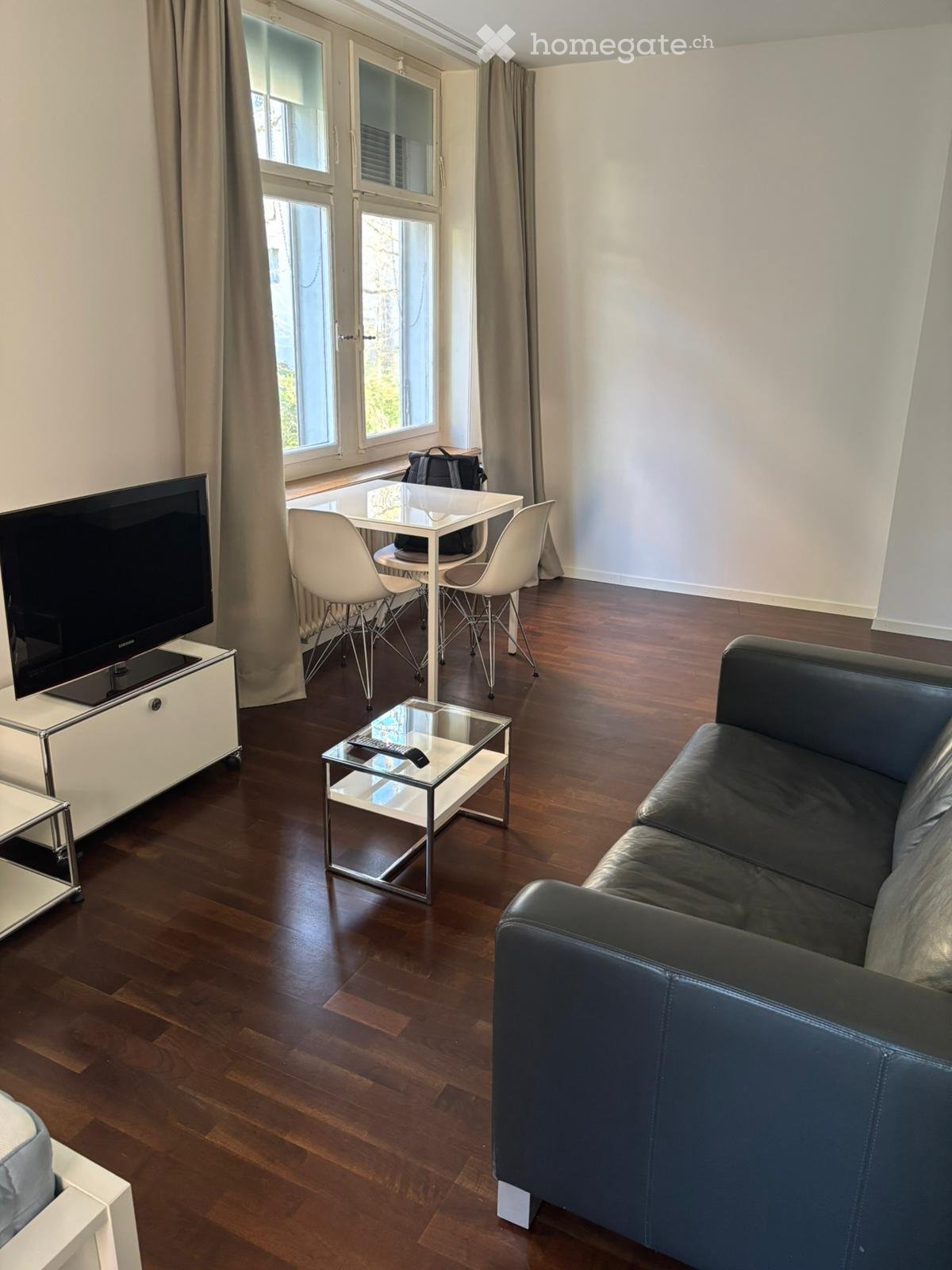 Modern living room with hardwood flooring, large windows, a black leather sofa, a glass coffee table, a TV on a white stand, and a small dining area with white chairs and a table.