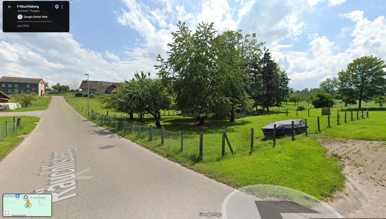 Rural outdoor scene with a paved driveway leading to a house, surrounded by green fields, trees, and a fenced area with a covered boat.