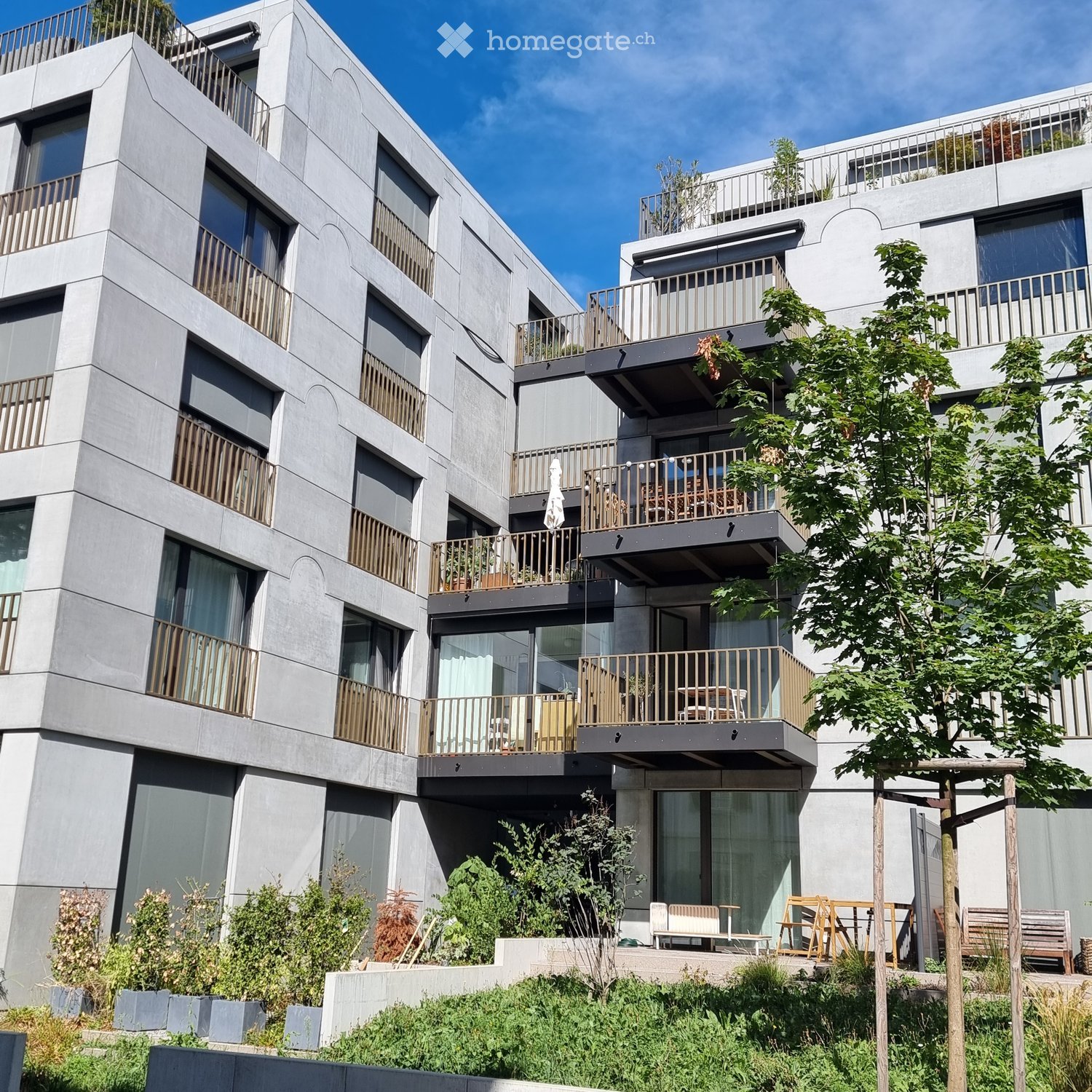 Modern multi-story residential building with grey concrete facade, large windows, and balconies with wooden railings. Greenery and seating areas are visible at the base.