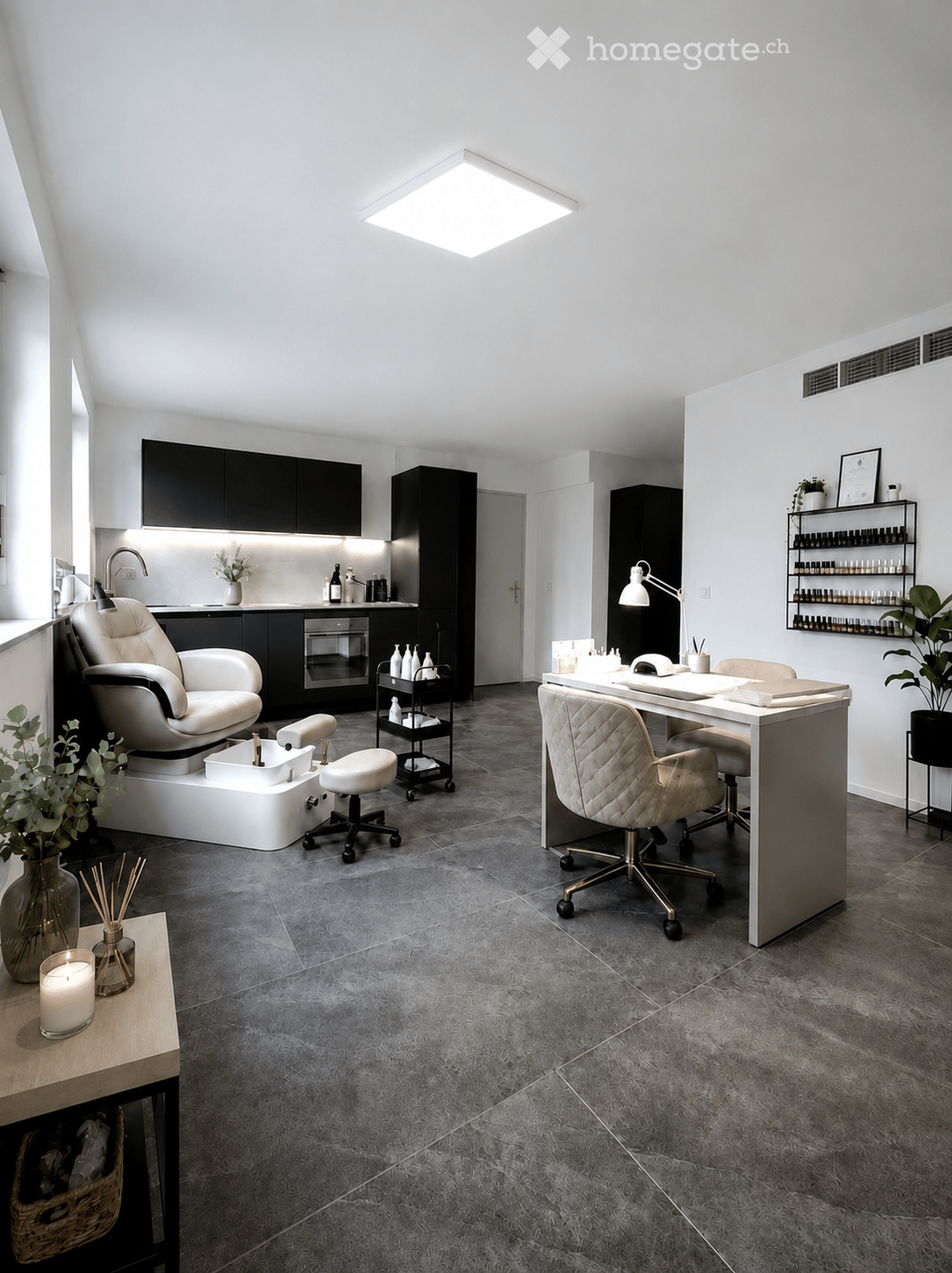 Modern, well-lit salon or spa room with a grey tiled floor, white walls, and black cabinets. Features include a large white massage chair, a white desk with a chair, a small sink area, a rolling cart with bottles, and decorative plants and candles.