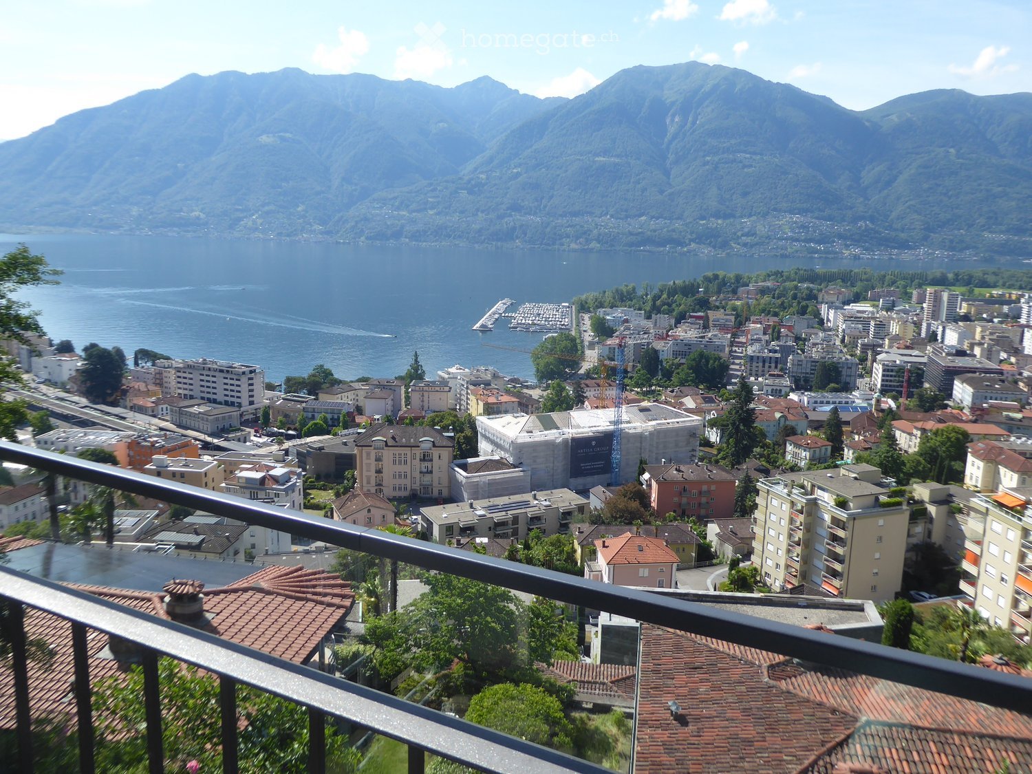Panoramic view from a balcony overlooking a cityscape, lake, and mountains.