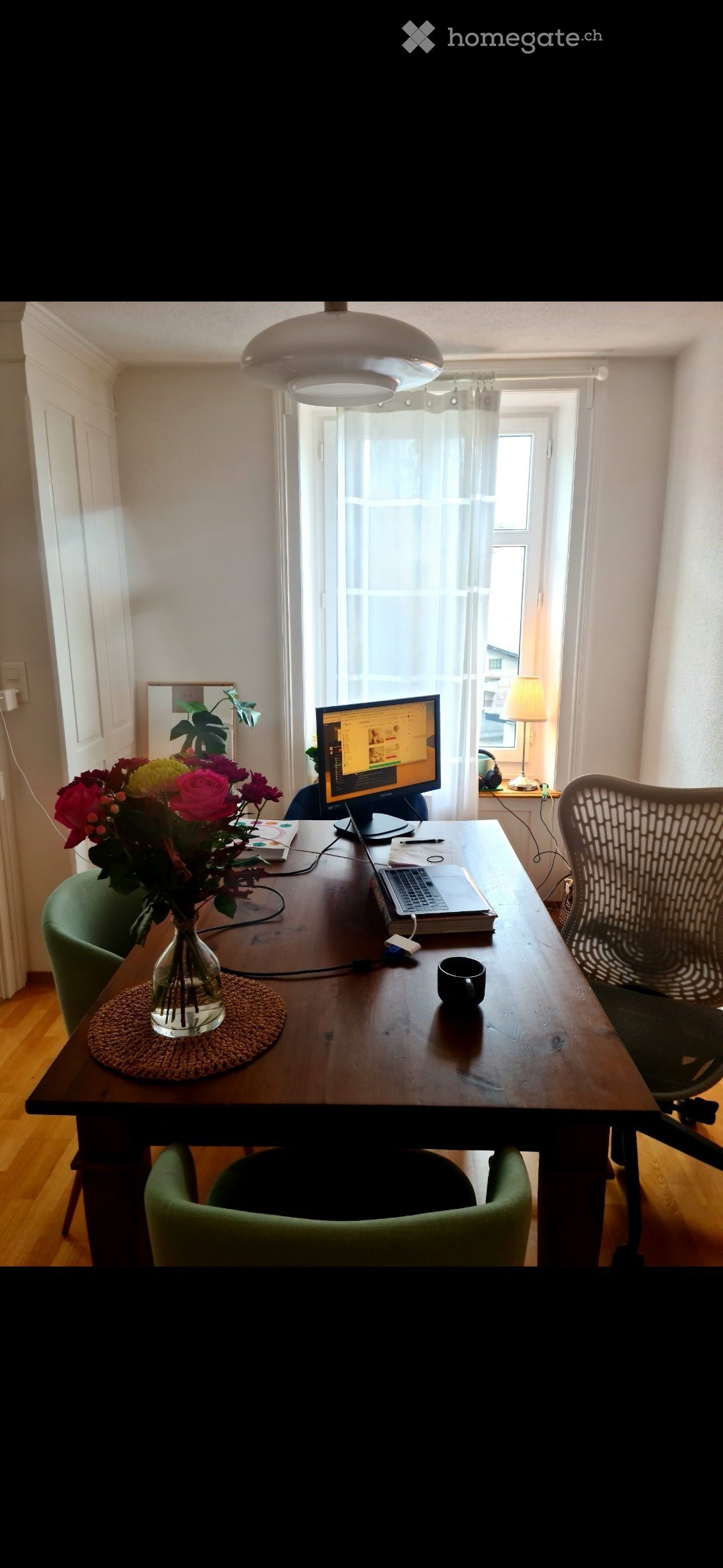 A well-lit room with a wooden table, green chairs, a laptop, a vase with flowers, and a lamp on a side table.
