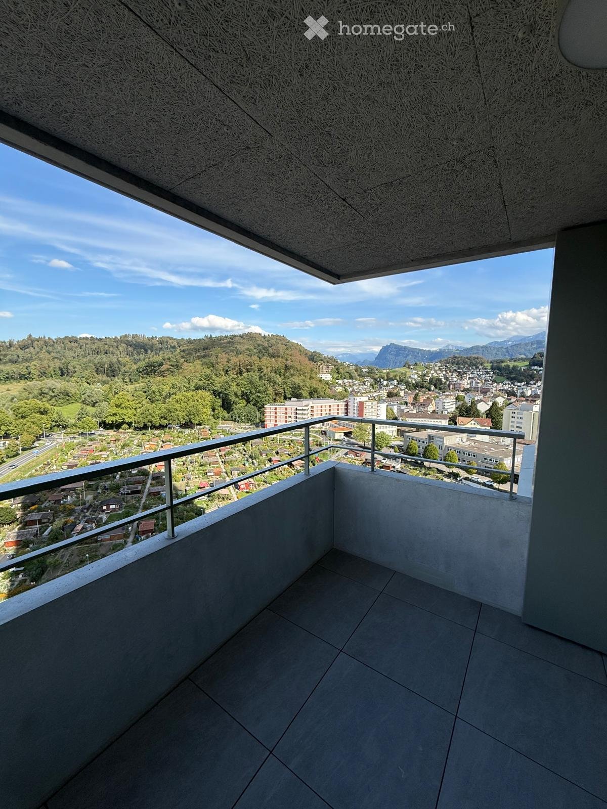 Modern balcony with grey tiled floor, metal railing, and expansive view of a town and surrounding mountains.