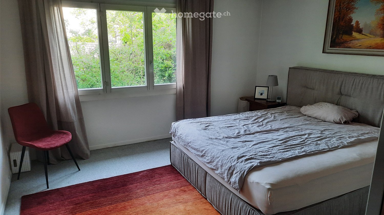 Bedroom with a double bed, nightstand, lamp, chair, and a picture frame on the wall. Large windows with curtains provide natural light.
