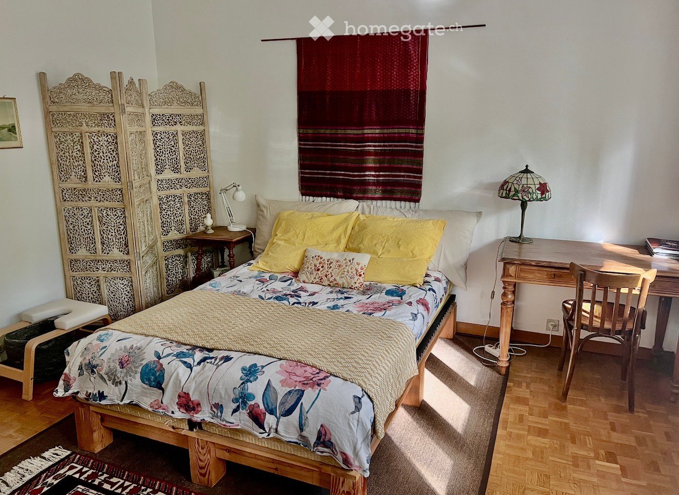 Cozy bedroom with a wooden bed, yellow pillows, floral duvet, nightstand with lamp, desk with chair, and decorative screen.