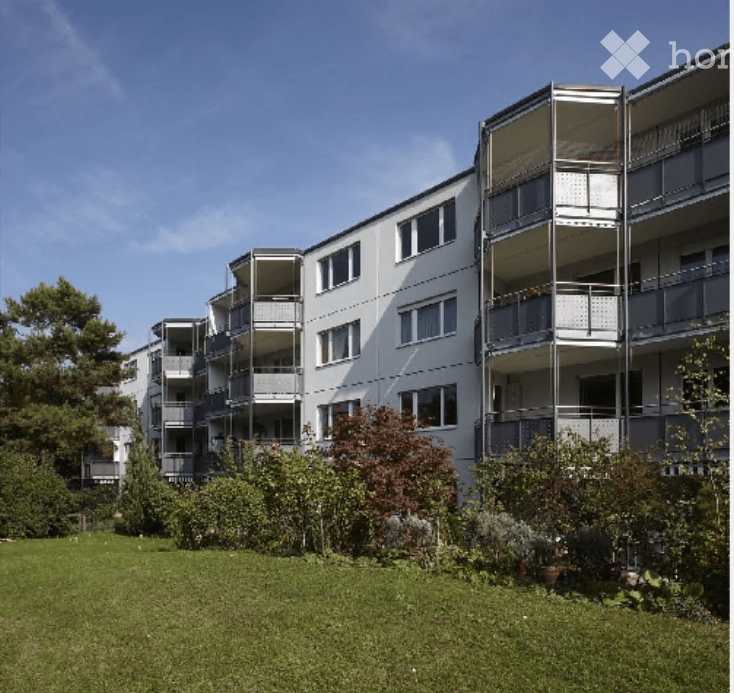 Modern multi-story apartment building with white facade, multiple balconies, and surrounding greenery.