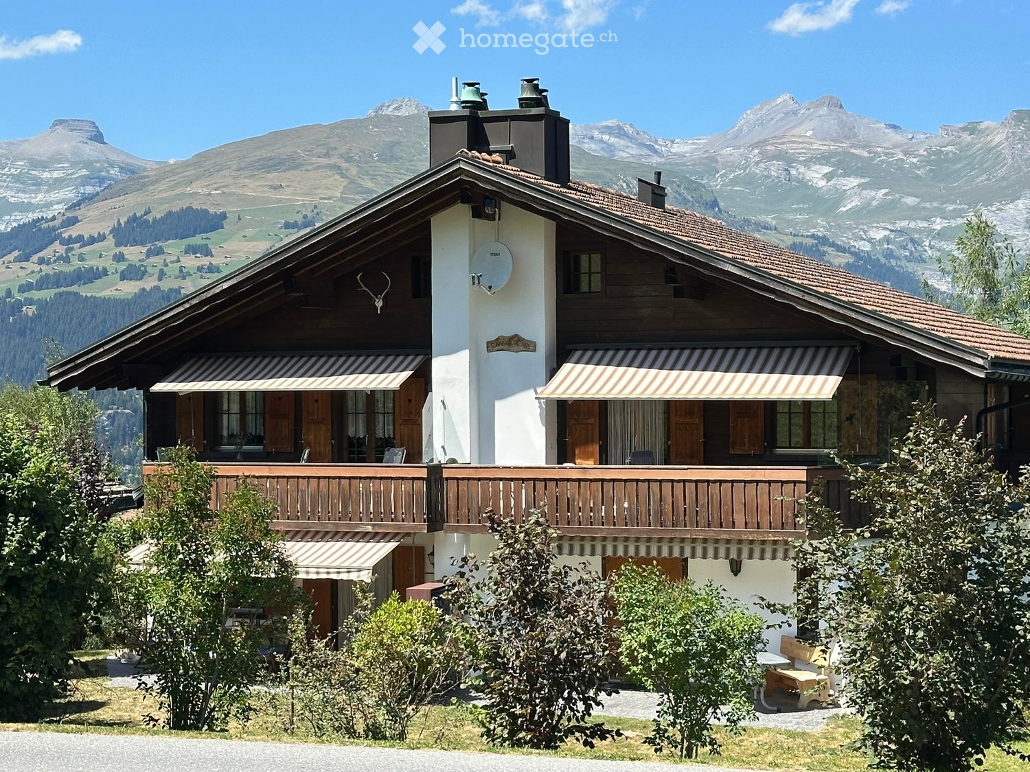 Two-story wooden house with white walls, striped awnings, balconies, and a scenic mountain backdrop.