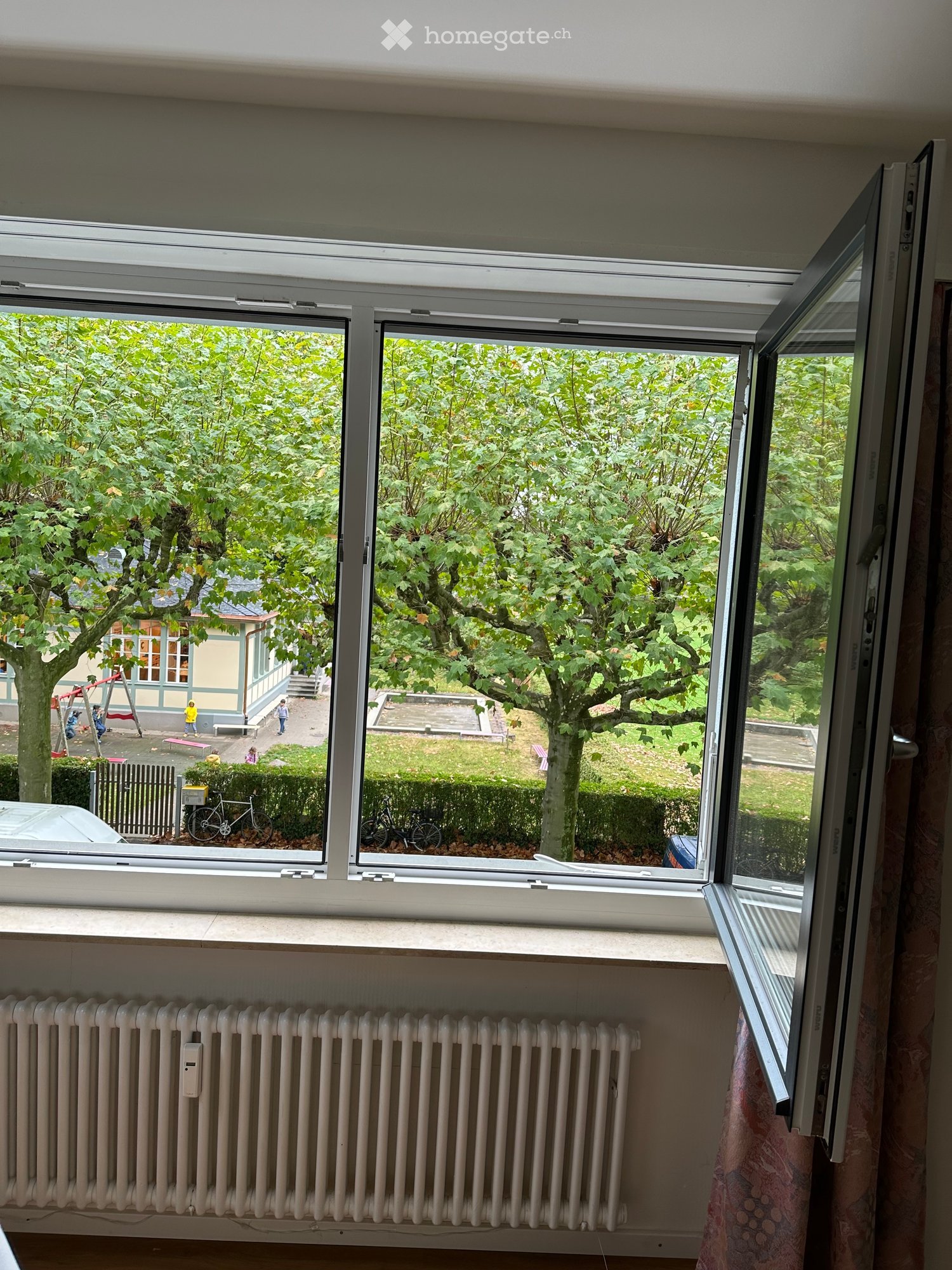 An unfurnished room with large windows offering a view of a park-like area with trees, a playground, and a house in the background. The room features a radiator and a partially visible curtain.