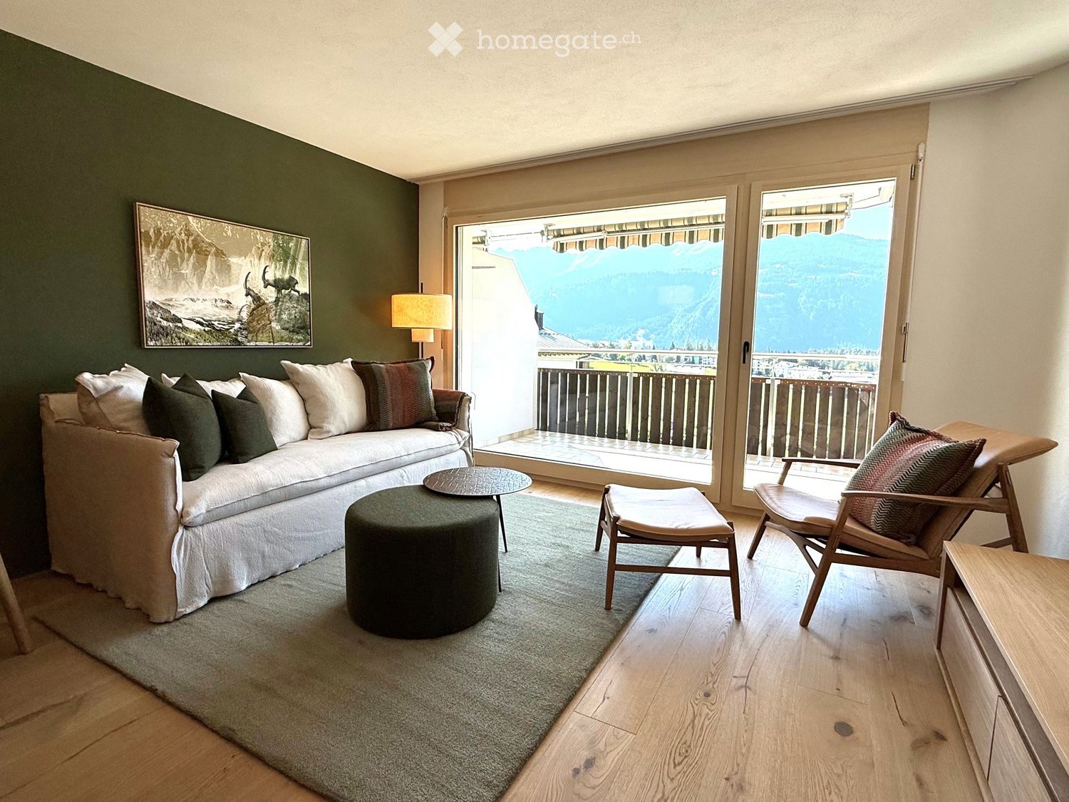 Modern living room with a large window offering a scenic mountain view, featuring a white sofa, coffee table, round stool, two chairs, a lamp, and a picture frame on the wall.