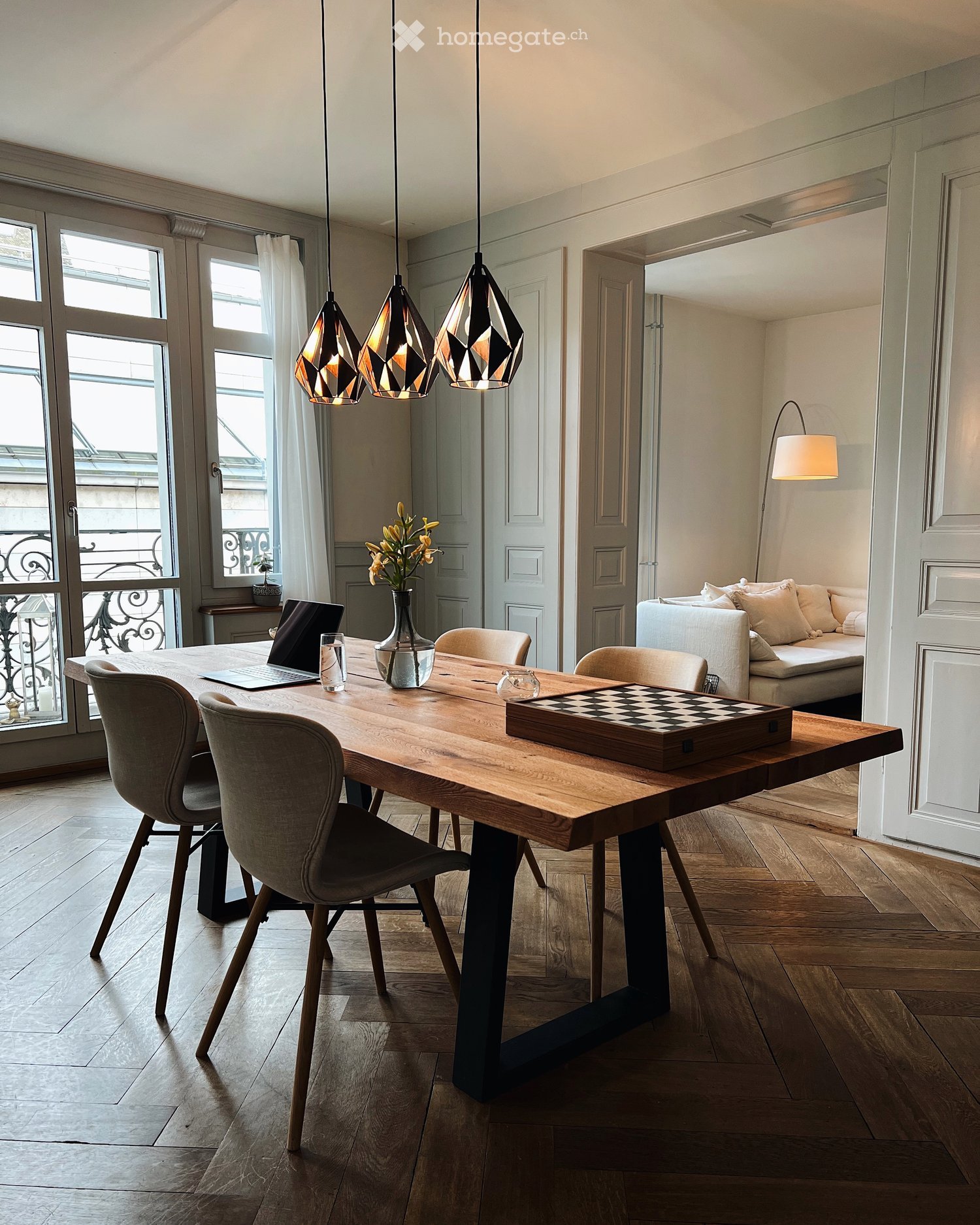 Modern dining area with a wooden table, chairs, laptop, vase with flowers, and a sofa in the background.