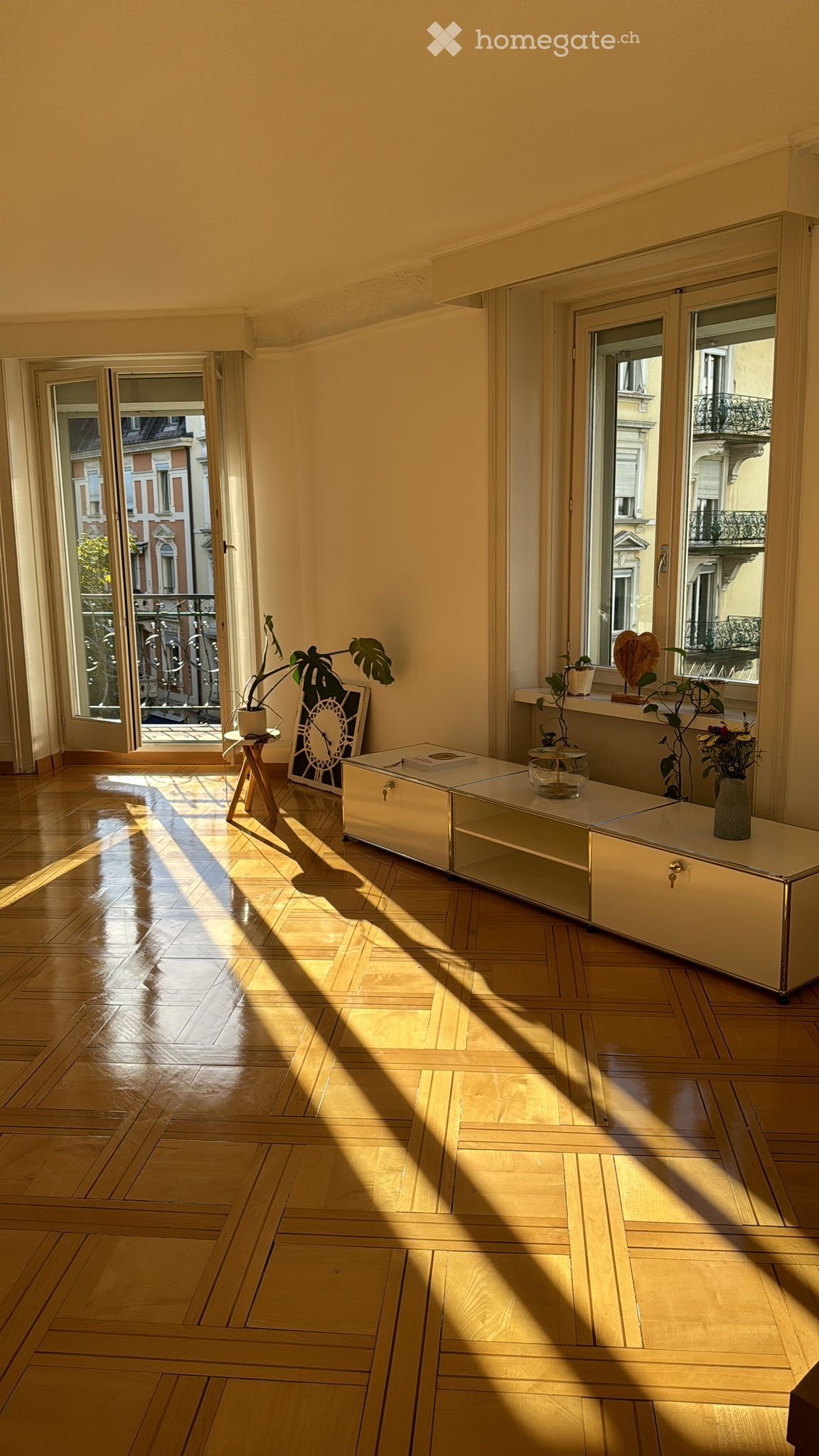 Bright, sunlit room with wooden parquet flooring, large windows, and a balcony. Contains a small table with a plant, a cabinet, and decorative items.