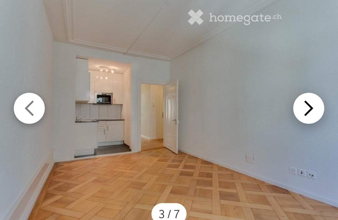 Empty room with light blue walls and wooden herringbone flooring. An open doorway leads to a small kitchen area with white cabinets and a microwave.