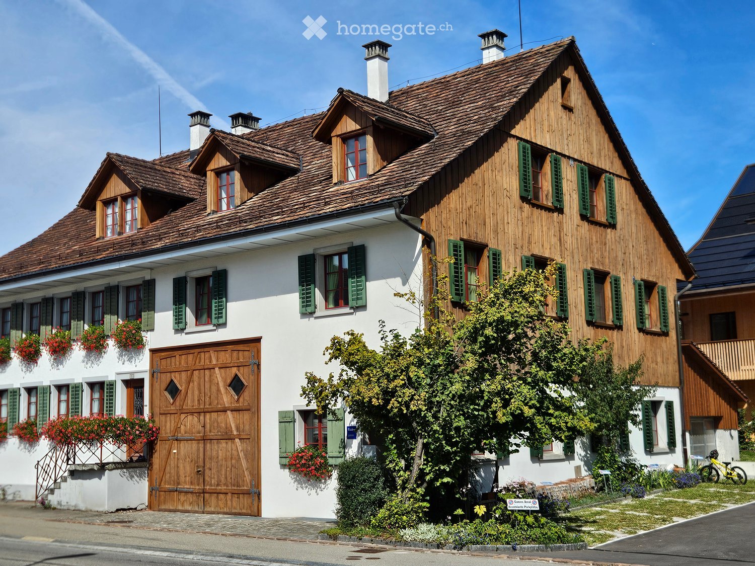 Traditional two-story house with a wooden facade, tiled roof, multiple chimneys, green shutters, and flower boxes on the windowsills.
