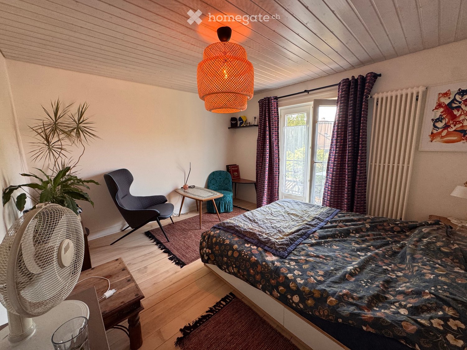 Cozy bedroom with a wooden bed, colorful quilt, a wicker fan, a modern chair, a small table, and a large plant near the window.