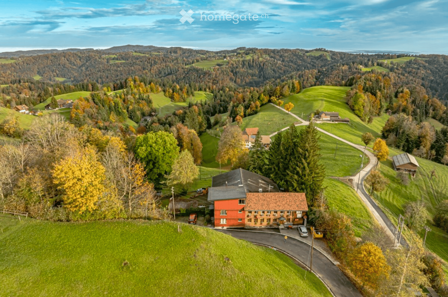 Aerial view of a rural area with a red house, surrounded by green fields, trees, and a winding road.