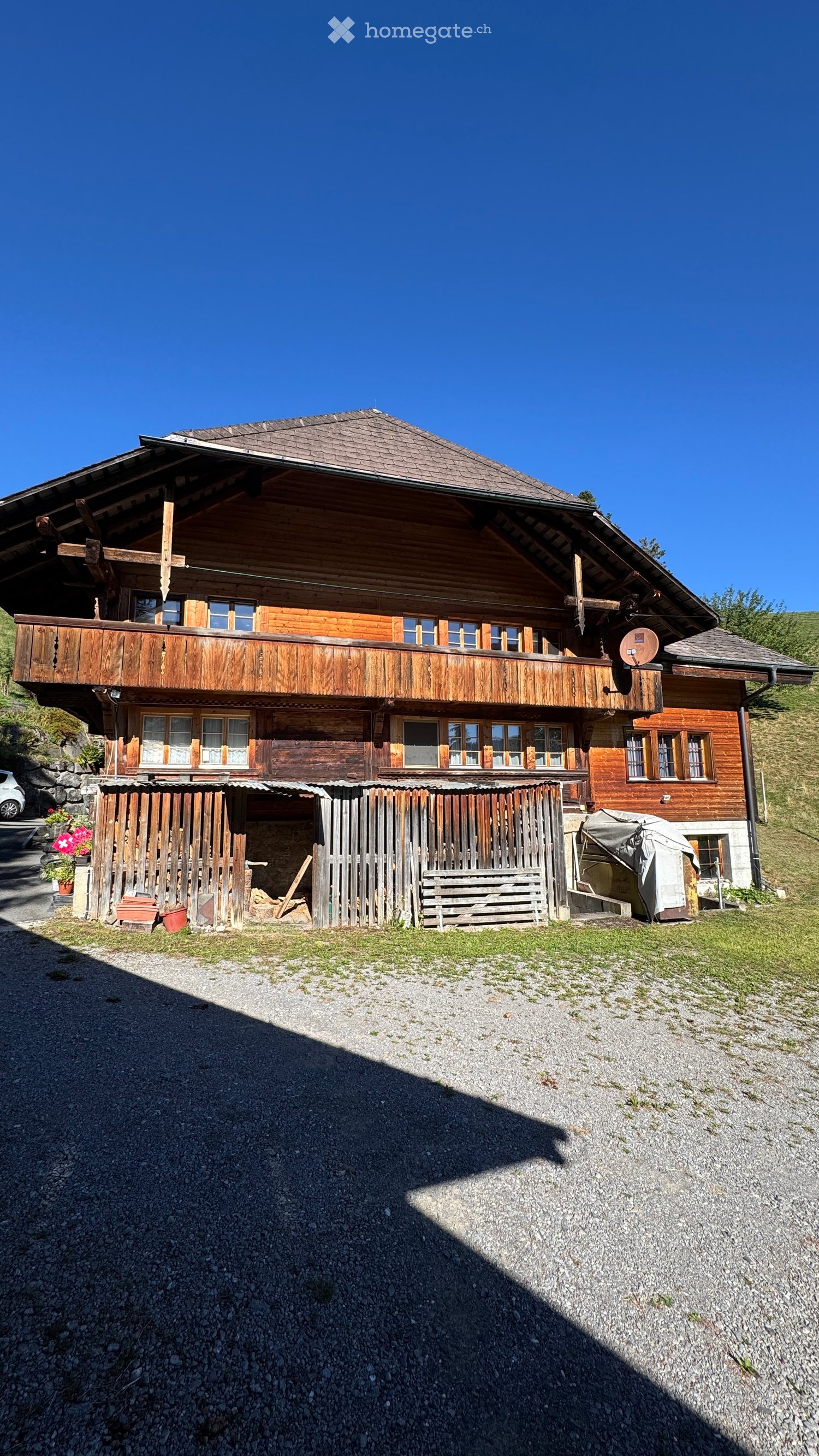 A two-story wooden house with a large balcony, multiple windows, and a wooden fence in front. The house has a traditional alpine design with a steeply pitched roof and is surrounded by a gravel driveway.