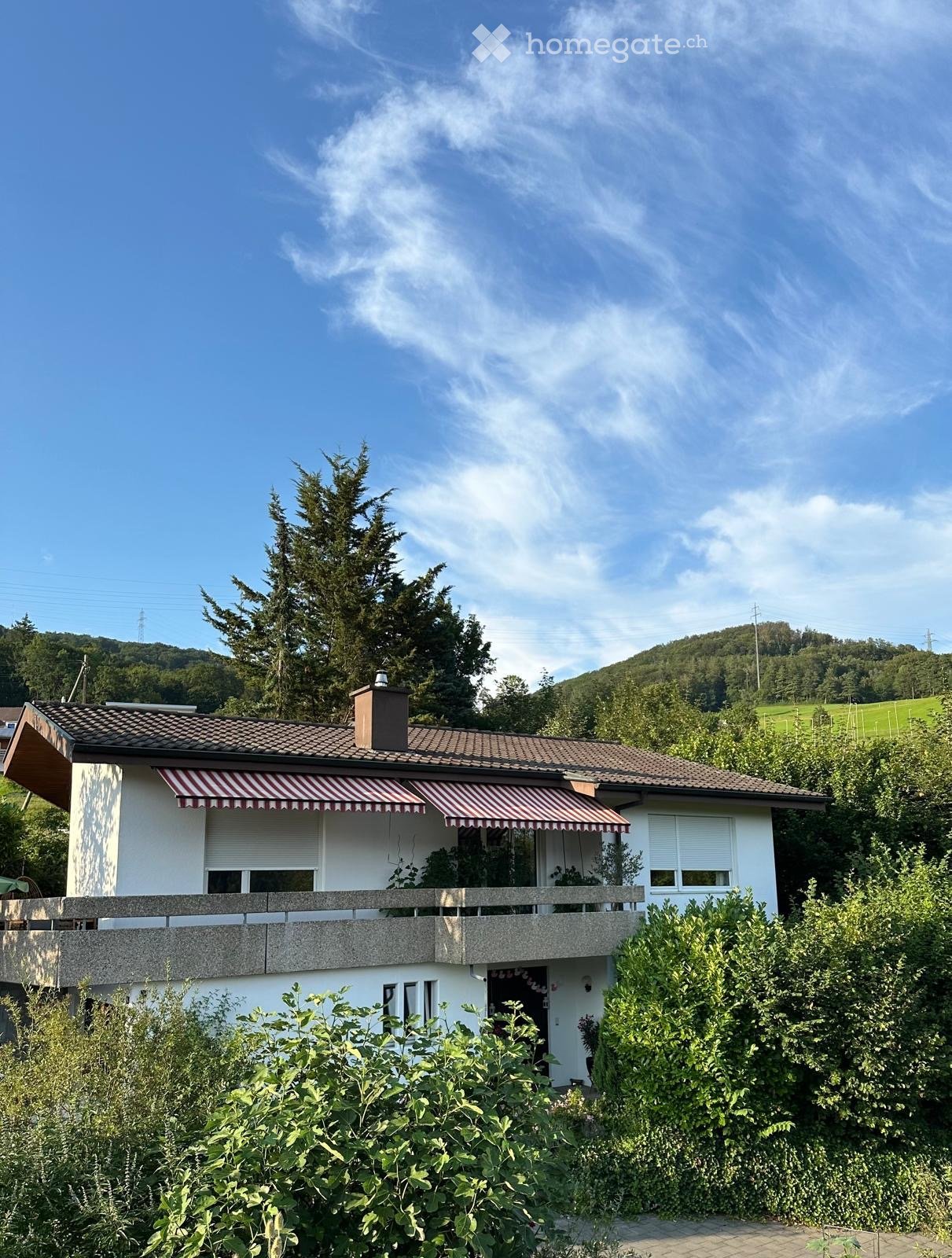 Two-story house with a tiled roof, white exterior, and striped awnings over the windows. The house features a balcony on the upper level and is surrounded by greenery and trees.
