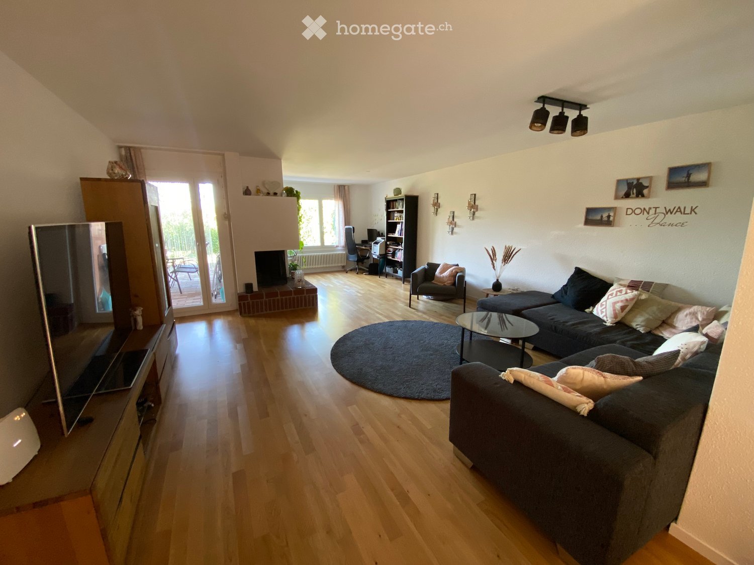 Bright living room with wooden flooring, large windows, a grey sectional sofa, coffee table, armchair, TV stand, bookshelf, and a balcony access.
