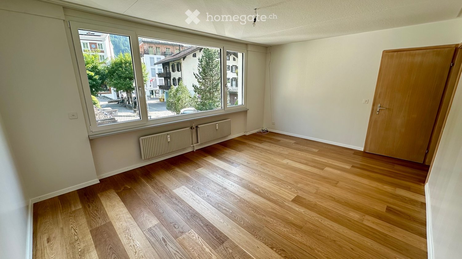 Empty room with wooden flooring, large windows, radiator, and a wooden door.