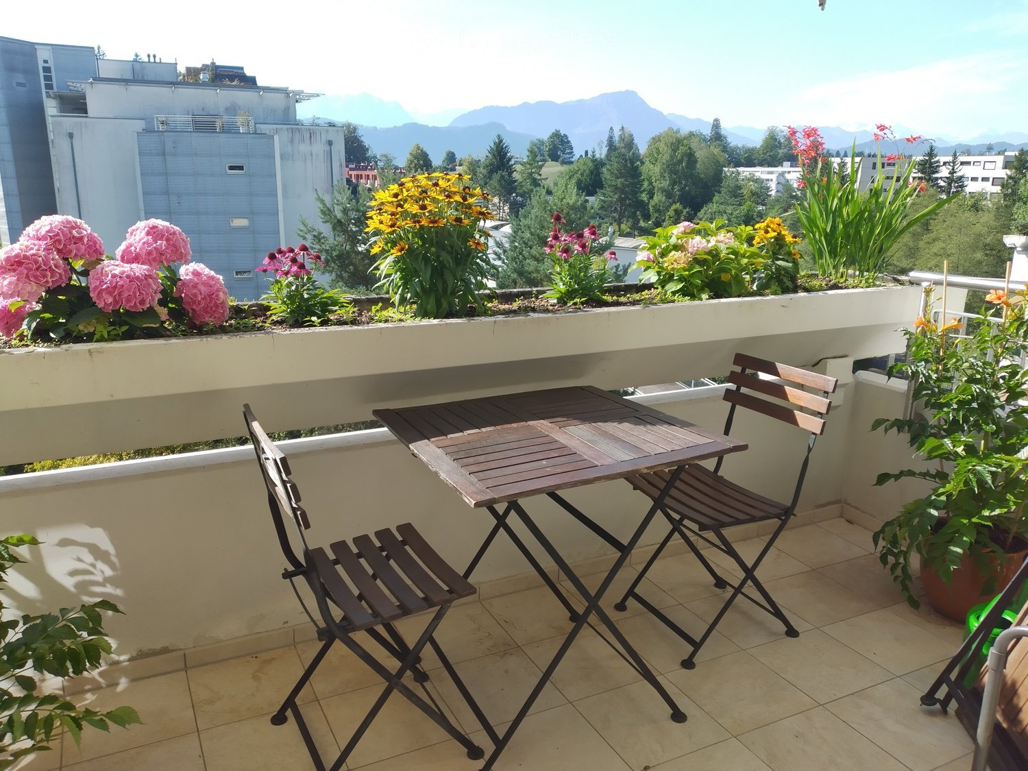 Balcony with a small wooden table and two folding chairs, surrounded by potted flowers and a scenic mountain view.