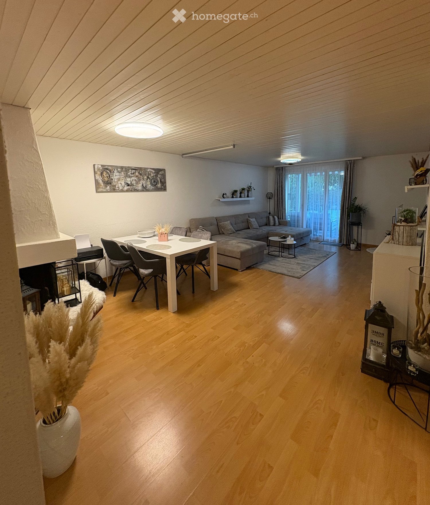 A modern living room with wooden flooring, a sectional sofa, a coffee table, a dining table with chairs, and a fireplace. The room features large windows with curtains and decorative elements like vases and artwork.