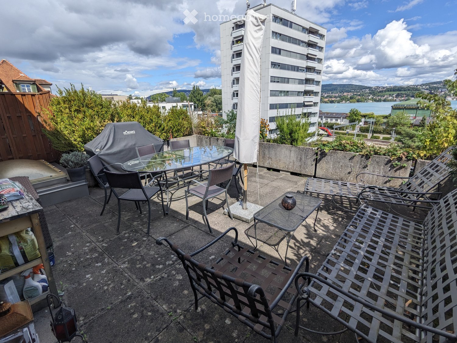 Balcony with a glass table, chairs, umbrella, and metal benches, overlooking a cityscape with a river and buildings.