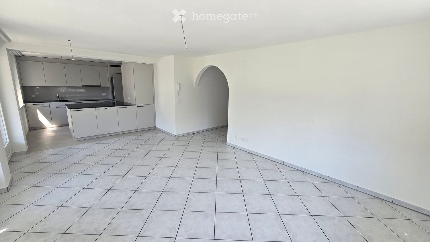 Bright, open-plan space with tiled flooring, white kitchen cabinets, and a sink area. The room features an arched doorway leading to another area.