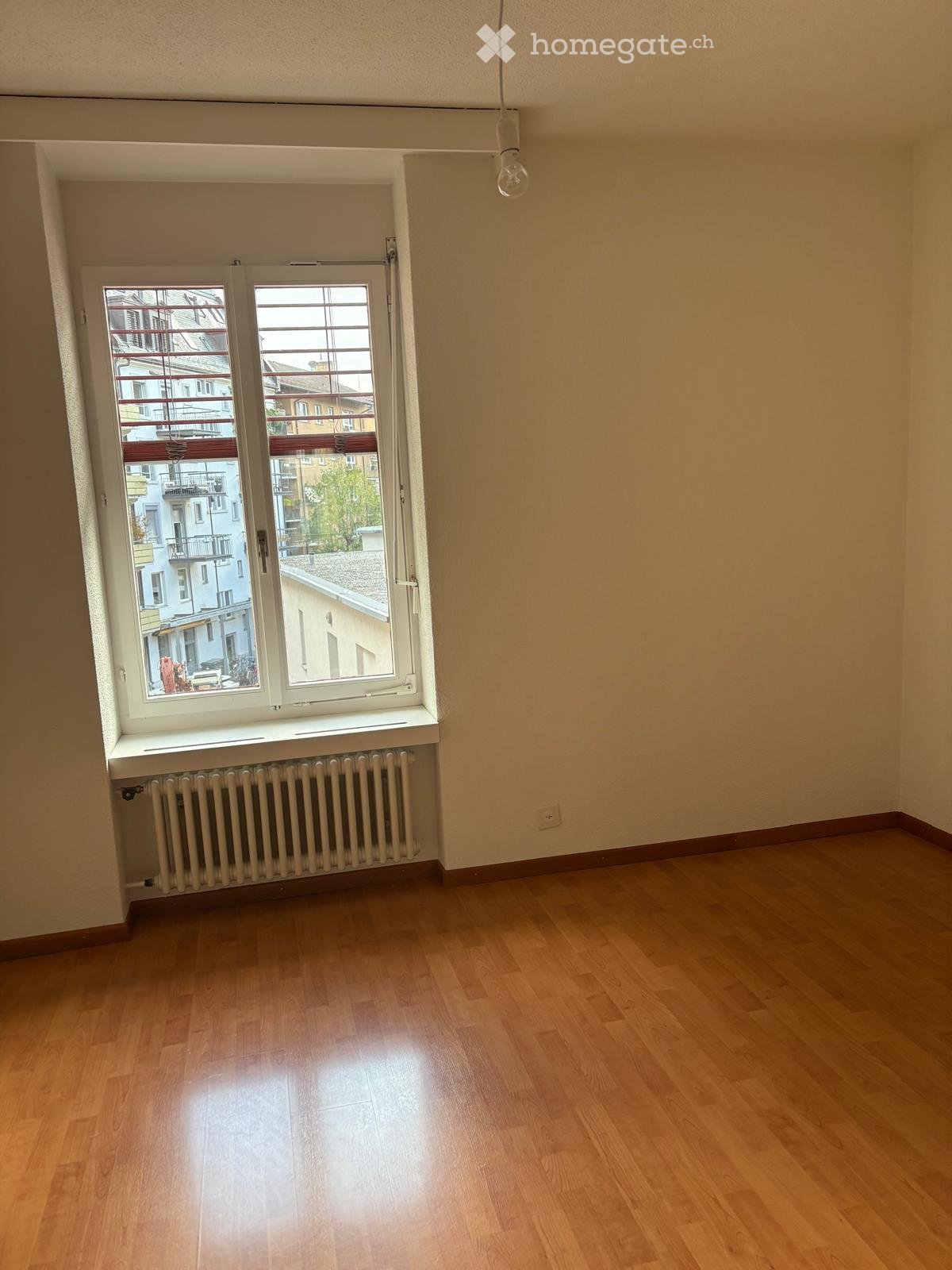 Empty room with wooden flooring, large window with radiator below, and visible cityscape outside.