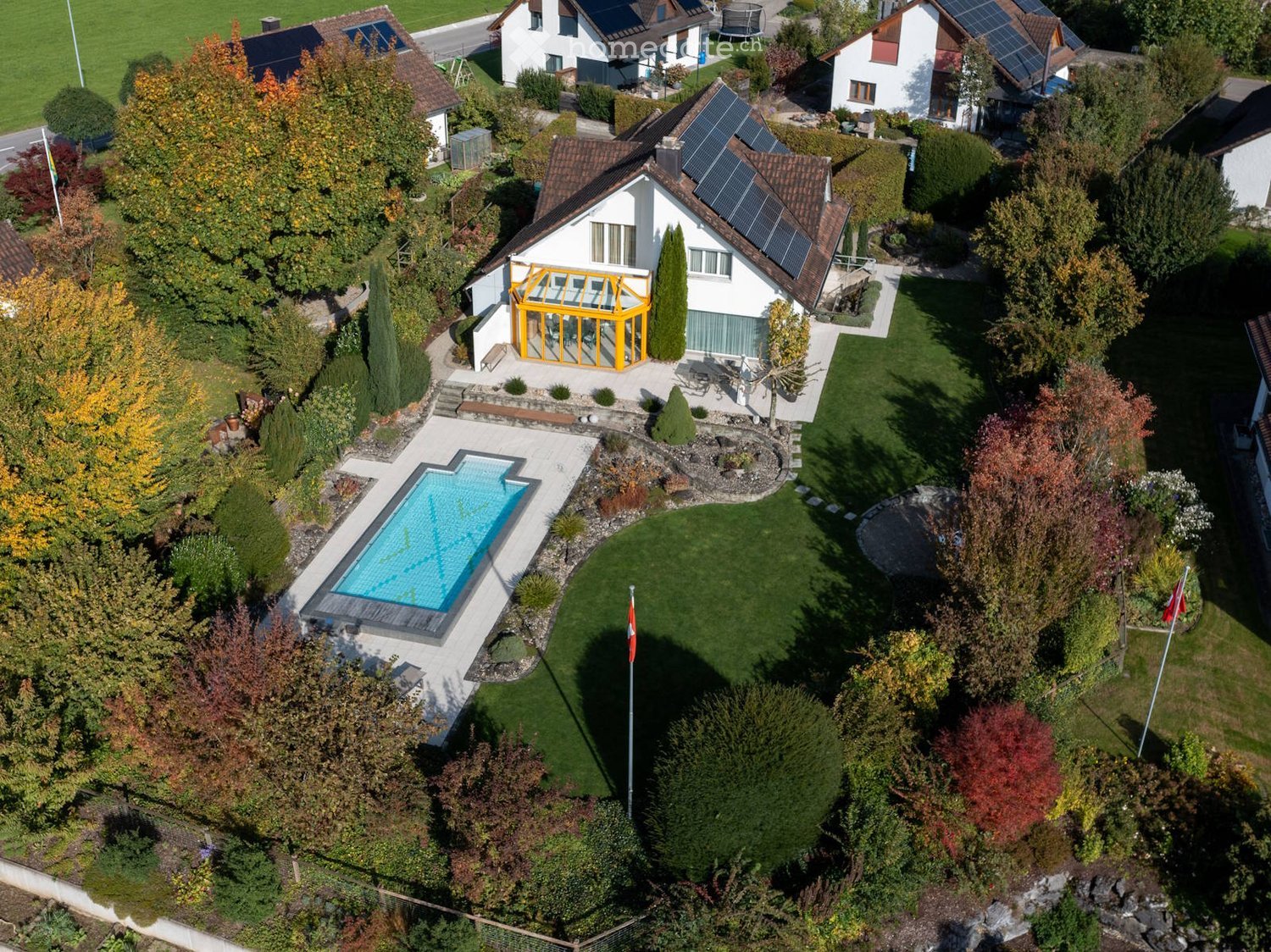 Aerial view of a house with a swimming pool, surrounded by a well-maintained garden, trees, and neighboring houses with solar panels on the roofs.