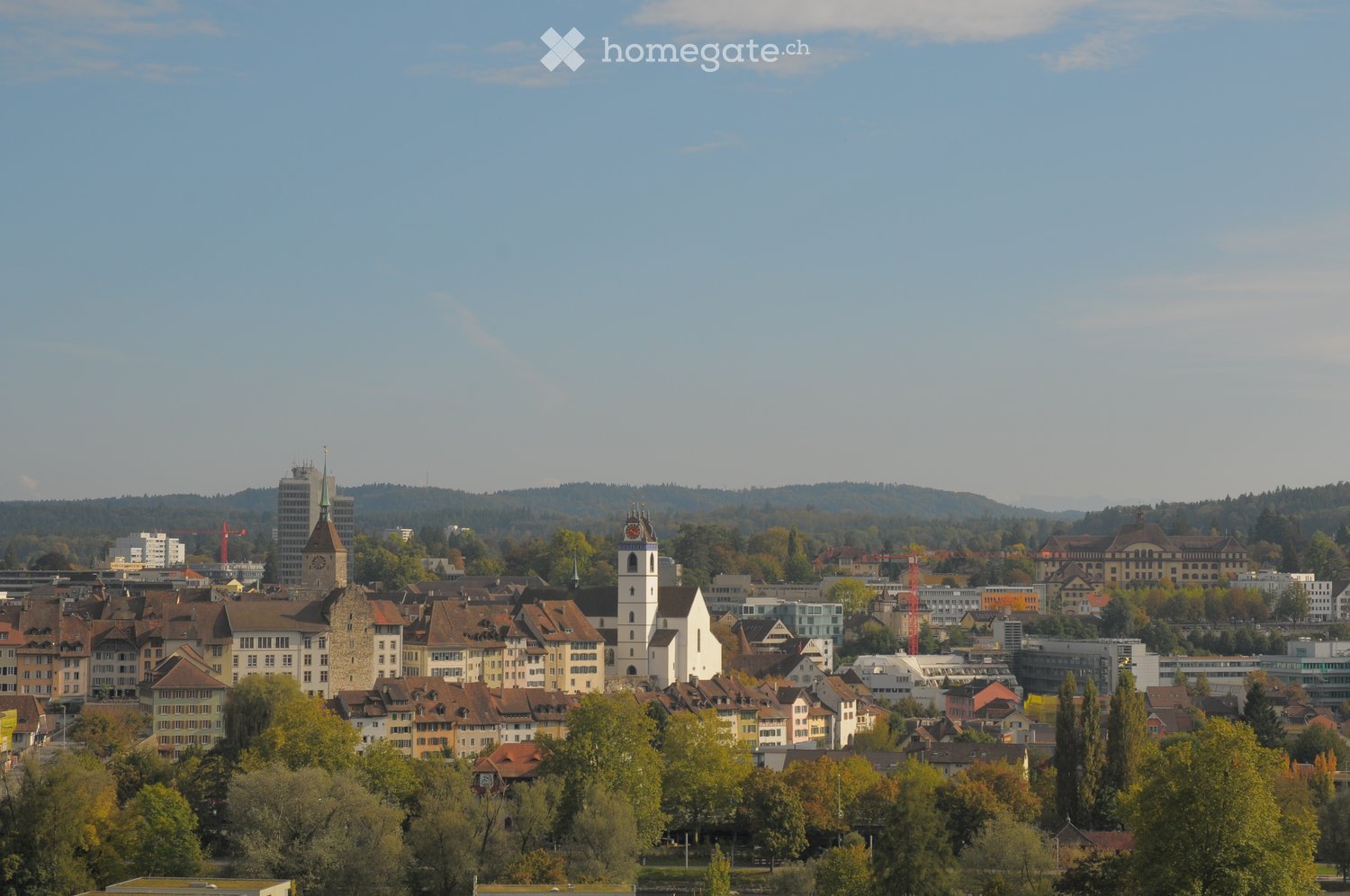 A panoramic view of a cityscape with a mix of historic and modern buildings, including a prominent church tower, surrounded by lush green trees and rolling hills in the background.
