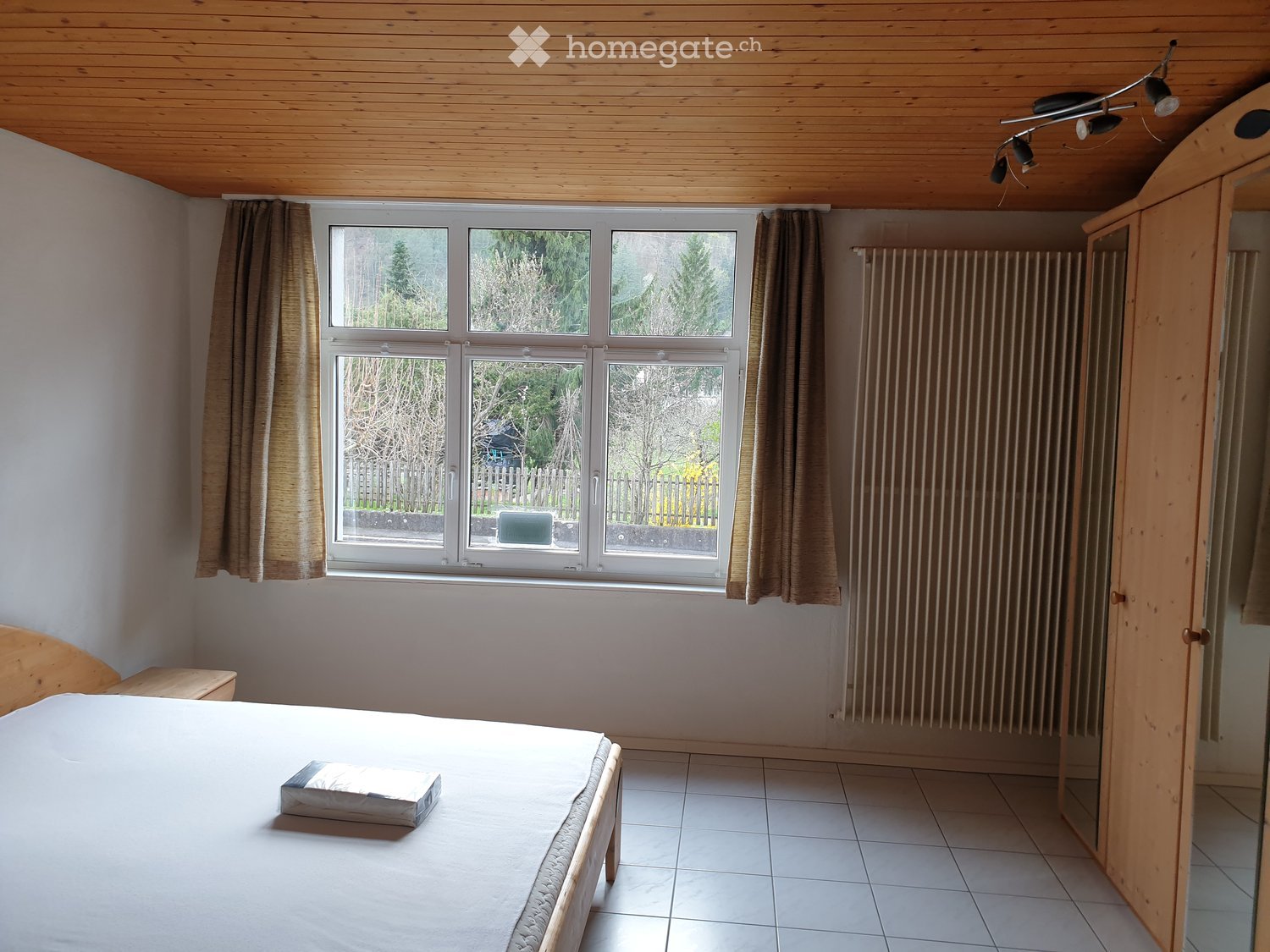 Bright bedroom with wooden ceiling, tiled floor, large windows with curtains, bed, radiator, and wardrobe.