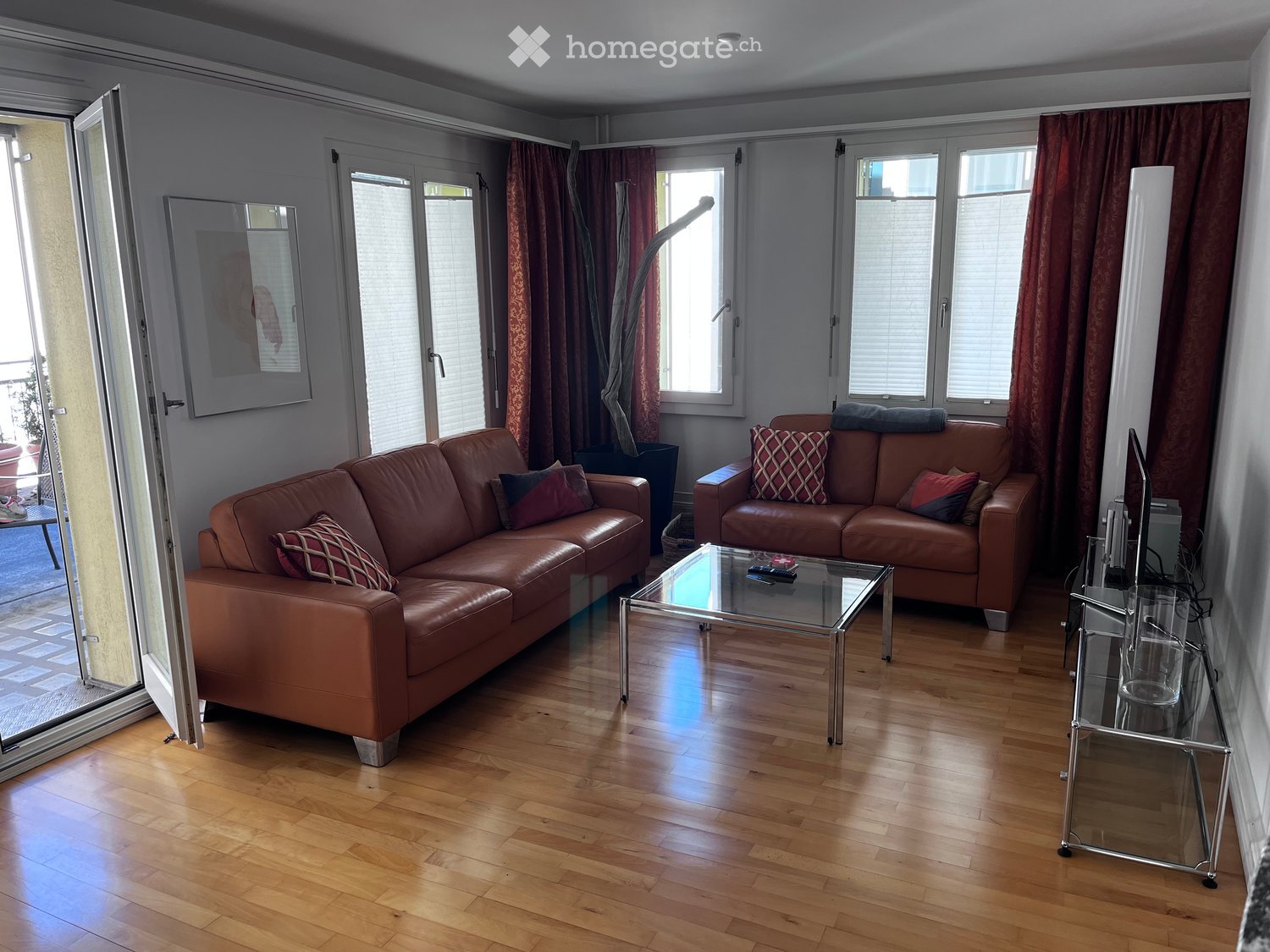 Modern living room with wooden flooring, large windows, and a balcony. Features two brown leather sofas, a glass coffee table, a TV stand, and a mirror on the wall.
