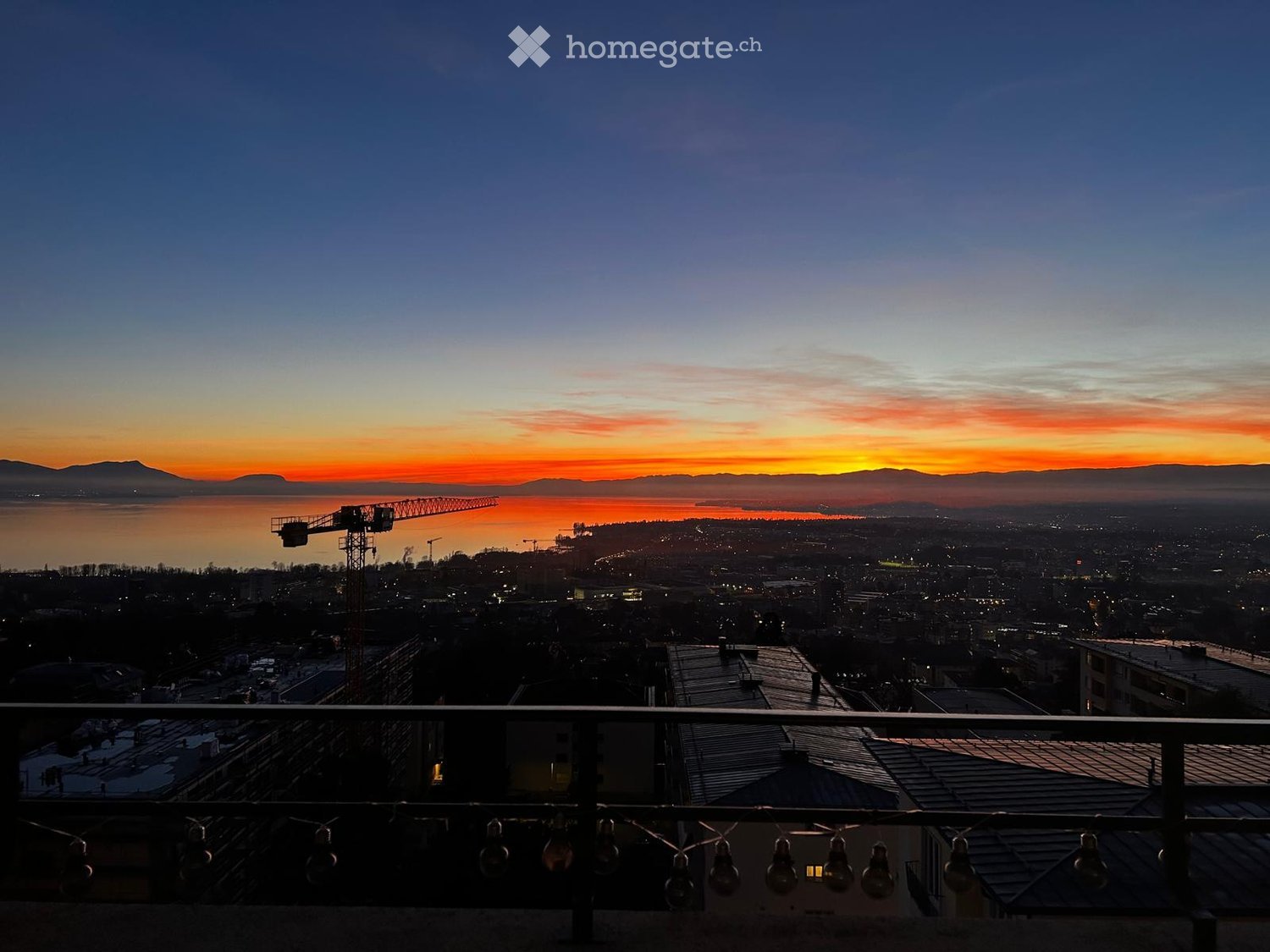 Balcony overlooking a cityscape at sunset, with a body of water and distant mountains visible.
