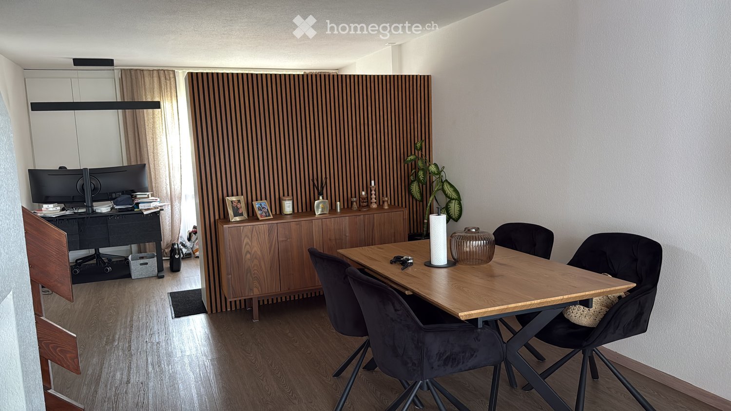 A modern, well-lit room with a wooden floor, a large wooden table surrounded by chairs, a desk with a computer screen, and a wooden cabinet with decorative items and a plant.