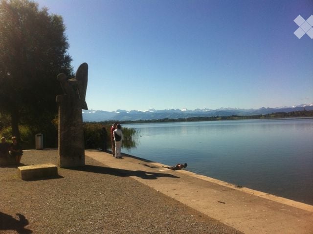 A lakeside promenade with a sculpture, a few people, and a scenic mountain view in the background.