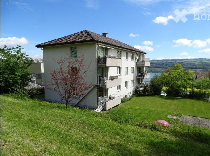 Multi-story residential building with balconies overlooking a scenic lake view, surrounded by green lawn and trees.