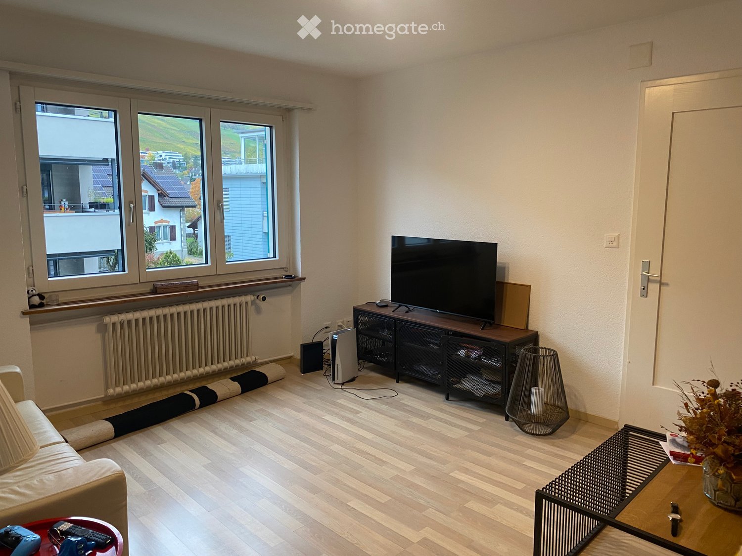 Modern living room with light wooden flooring, large windows offering a scenic view, a cream-colored sofa, a coffee table, a television on a stand, and a decorative floor lamp.
