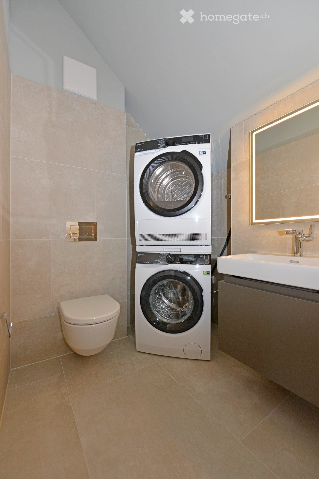 Modern bathroom with beige tiles, a toilet, a stacked washing machine and dryer, a sink with a mirror, and a sleek design.
