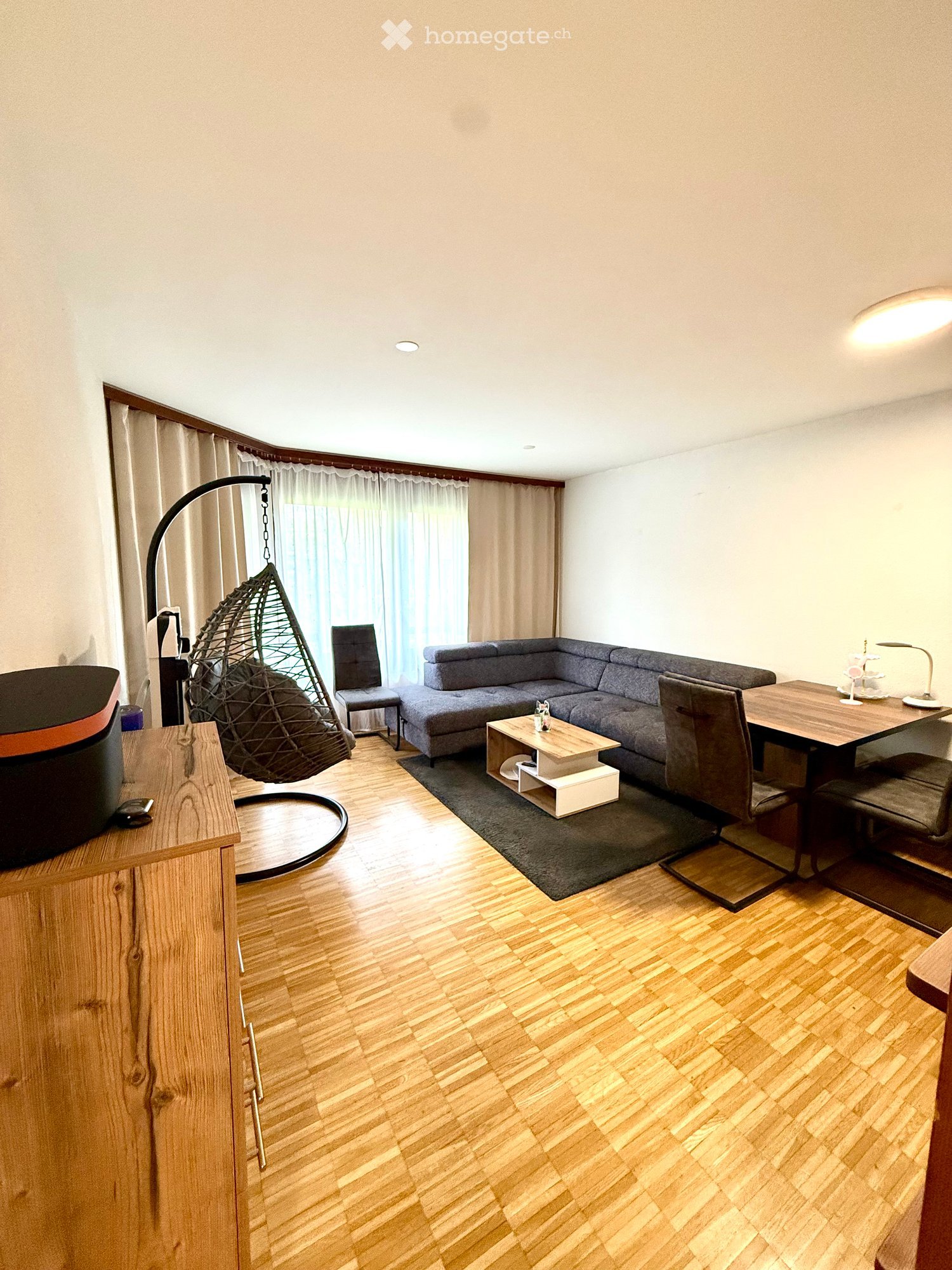 Modern living room with wooden flooring, grey sectional sofa, white coffee table, wicker hanging chair, dining table with chairs, and a wooden cabinet.