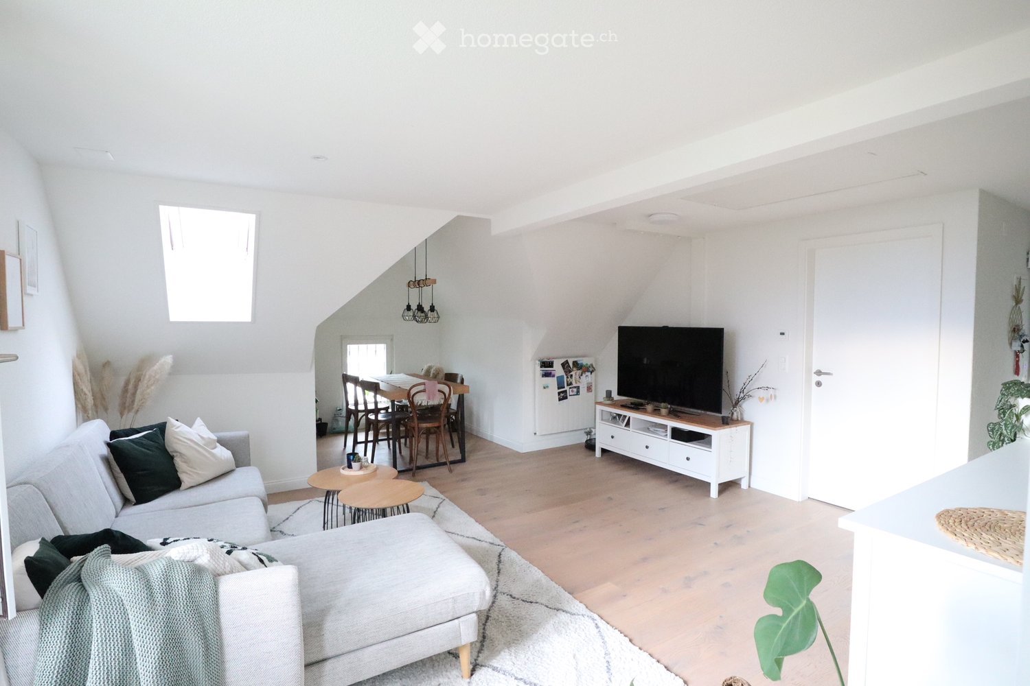 Bright, open-concept living and dining area with white walls, wooden flooring, and large windows. Features a grey sofa with pillows, an ottoman, a coffee table, a television on a stand, a dining table with chairs, and a refrigerator.