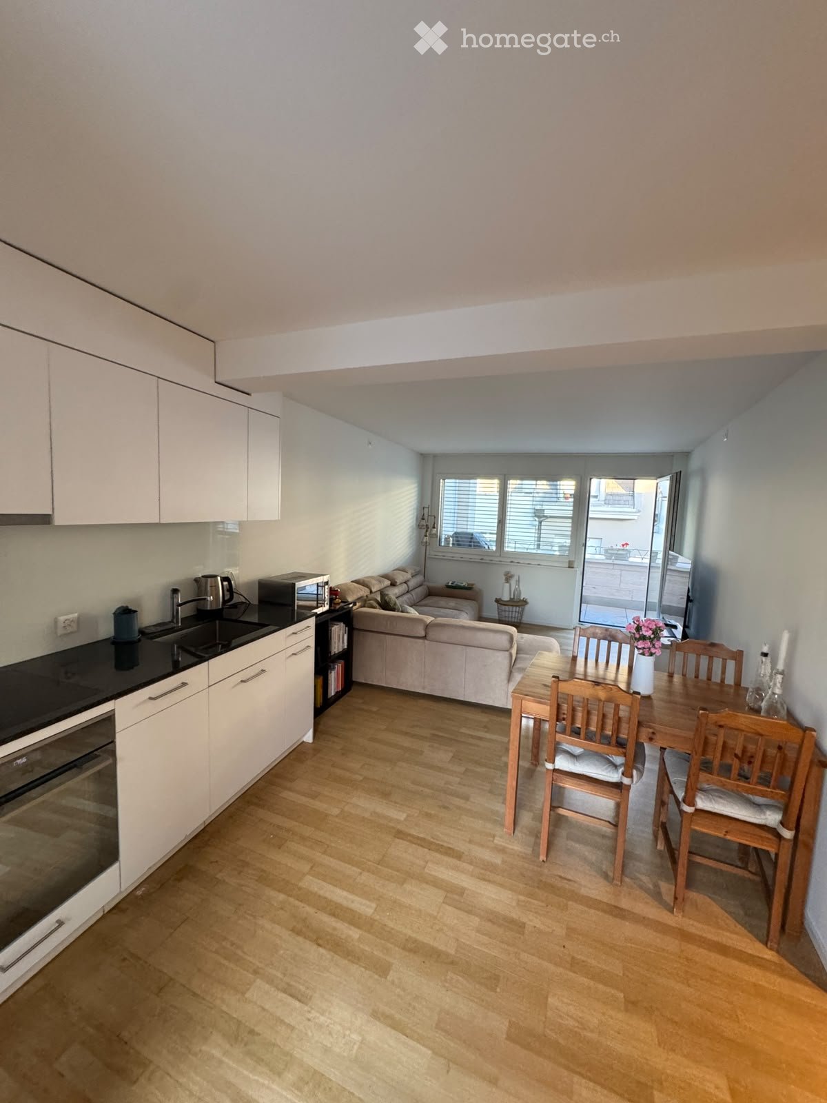 Open-plan living and dining area with a sofa, dining table, chairs, and a modern kitchen featuring white cabinets and stainless steel appliances.