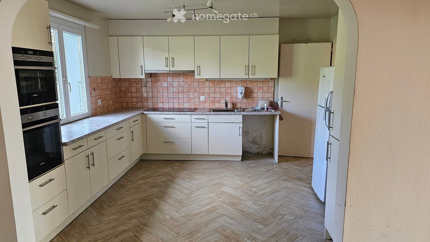 Modern kitchen with white cabinets, granite countertops, and wooden herringbone flooring. Features an oven, refrigerator, and sink.